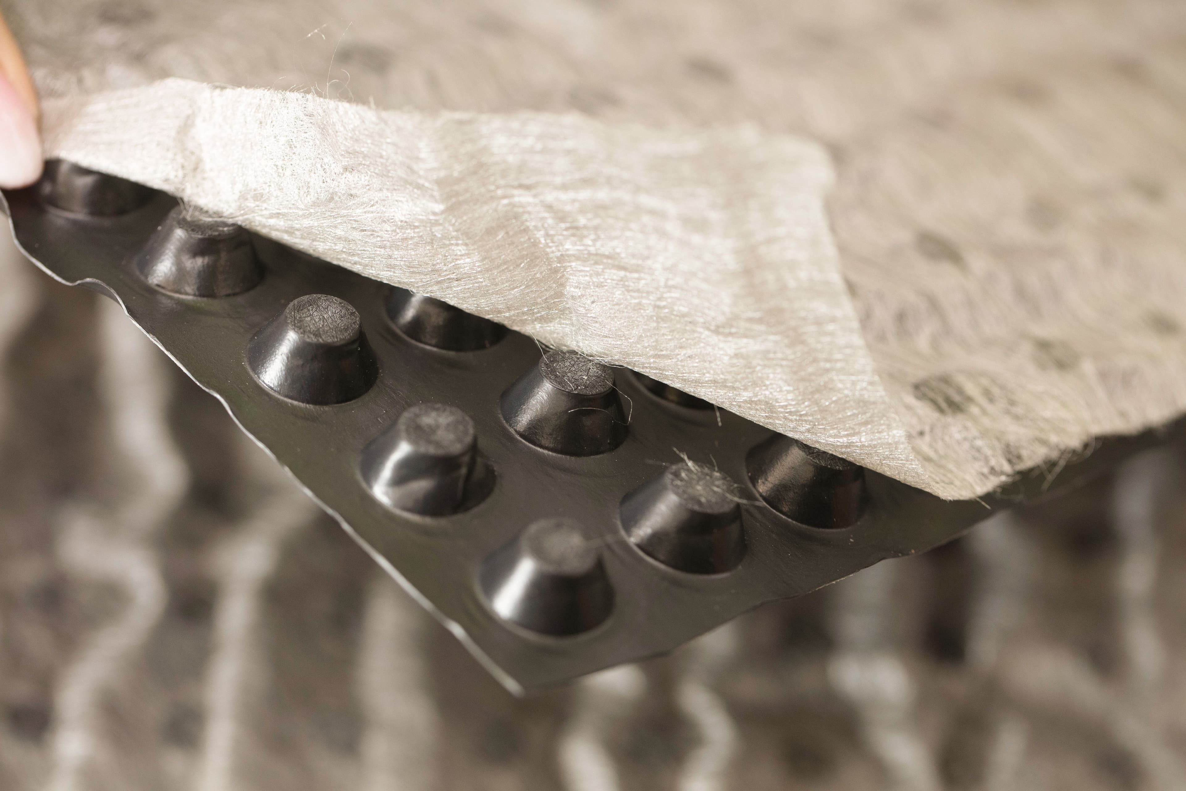 Black dimpled plastic drainage mat with a fibrous geotextile fabric layer partially peeled away.